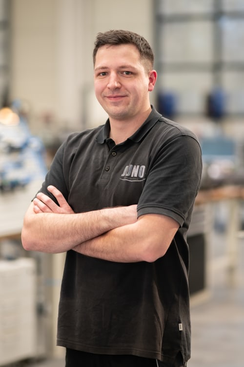 Lächelnder Mitarbeiter im schwarzen JUNO-Poloshirt, Arme verschränkt, in der Werkhalle