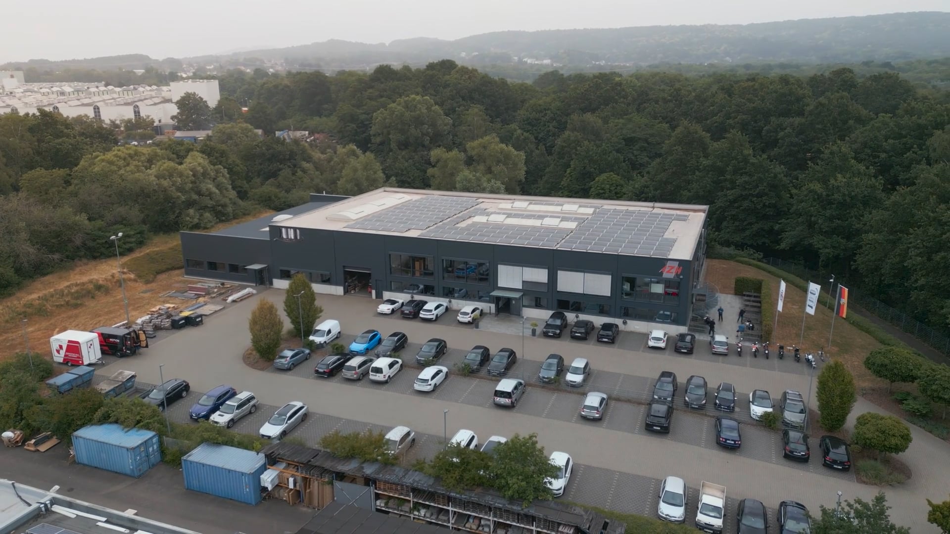 Luftaufnahme eines weißen Bürogebäudes mit Solaranlagen auf dem Dach, umgeben von Bäumen und einem Parkplatz mit Autos.