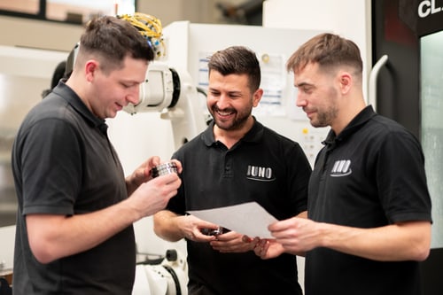 Drei Mitarbeiter in schwarzen JUNO-Poloshirts begutachten ein Metallteil und eine Zeichnung an einer Maschine