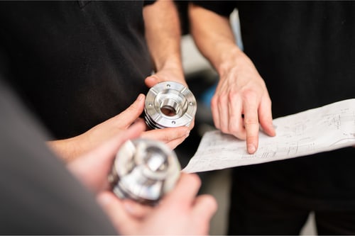 Hände halten zwei gefräste Metallteile und eine technische Zeichnung