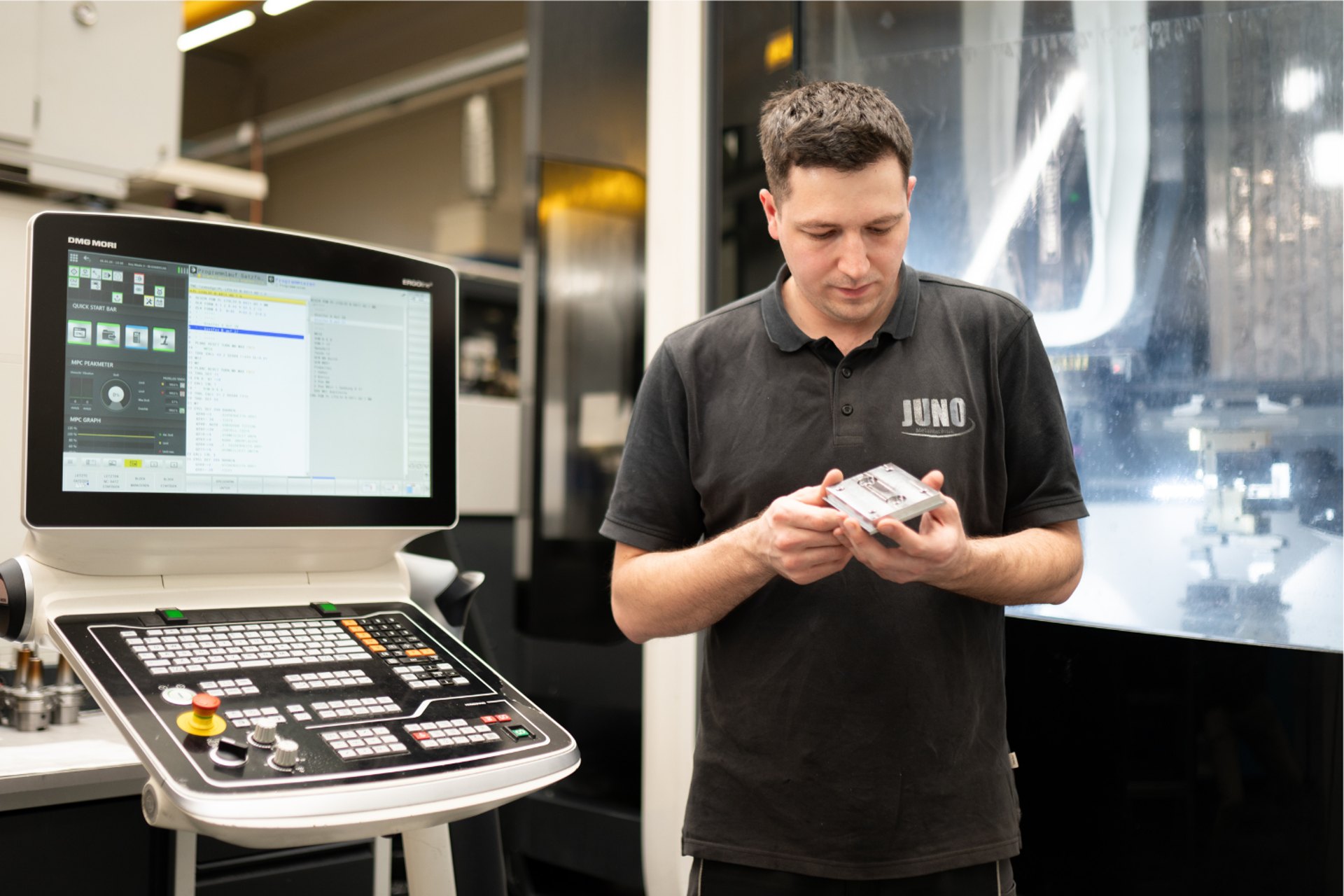 Mitarbeiter im JUNO-Poloshirt hält ein Metallteil vor dem Bedienpult einer CNC-Maschine